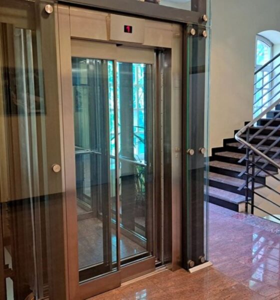 Na fotografiji se vidi moderno stakleno dizalo smješteno unutar stubišta zgrade. Dizalo ima prozirne stijenke i metalnu konstrukciju, što omogućuje pogled na unutrašnjost i okolni prostor. Uz dizalo se nalaze stepenice s tamnom metalnom ogradom koje vode na više katove. Pod je obložen sjajnim kamenom, a prostor djeluje uredno, svijetlo i suvremeno uređen.