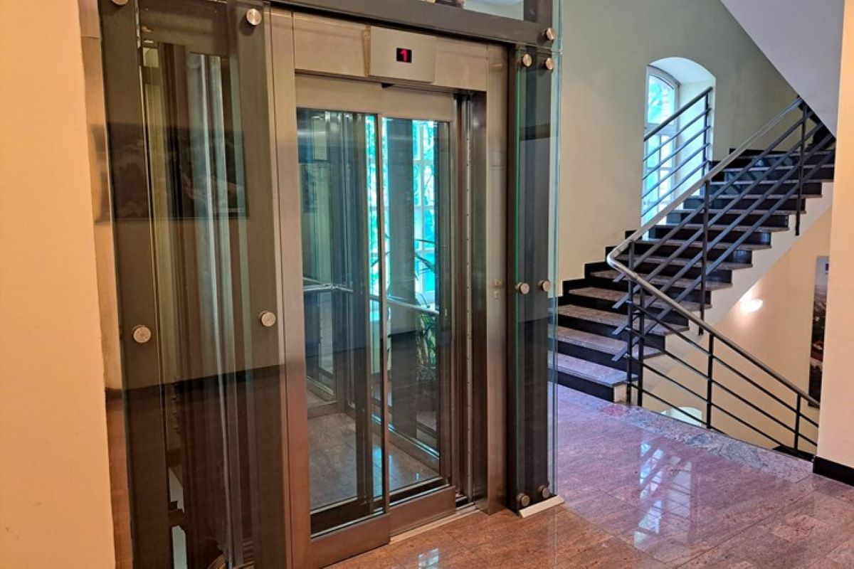 Na fotografiji se vidi moderno stakleno dizalo smješteno unutar stubišta zgrade. Dizalo ima prozirne stijenke i metalnu konstrukciju, što omogućuje pogled na unutrašnjost i okolni prostor. Uz dizalo se nalaze stepenice s tamnom metalnom ogradom koje vode na više katove. Pod je obložen sjajnim kamenom, a prostor djeluje uredno, svijetlo i suvremeno uređen.
