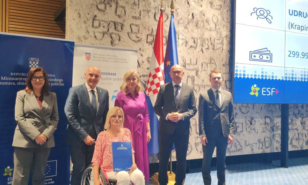 Na fotografiji se nalazi skupina od šest osoba u svečanom prostoru, vjerojatno tijekom službenog događanja ili dodjele ugovora. U prvom planu, u sredini, sjedi žena u invalidskim kolicima koja u rukama drži plavu mapu – najvjerojatnije potpisani ugovor ili projektni dokument. Oko nje stoje četiri muškarca i jedna žena, svi odjeveni u poslovnu, formalnu odjeću, što dodatno naglašava službeni karakter događaja. U pozadini se vide zastave Republike Hrvatske i Europske unije, kao i promotivni paneli s logotipima institucija i fondova. Na velikom ekranu s desne strane prikazan je naziv udruge i iznos projekta (299.999,14 eura), uz oznaku ESF+ fonda. Cjelokupna scena odaje dojam važnog i svečanog trenutka – potpisivanja i dodjele sredstava za projekt koji ima značajnu društvenu vrijednost, posebno u području podrške osobama s invaliditetom.