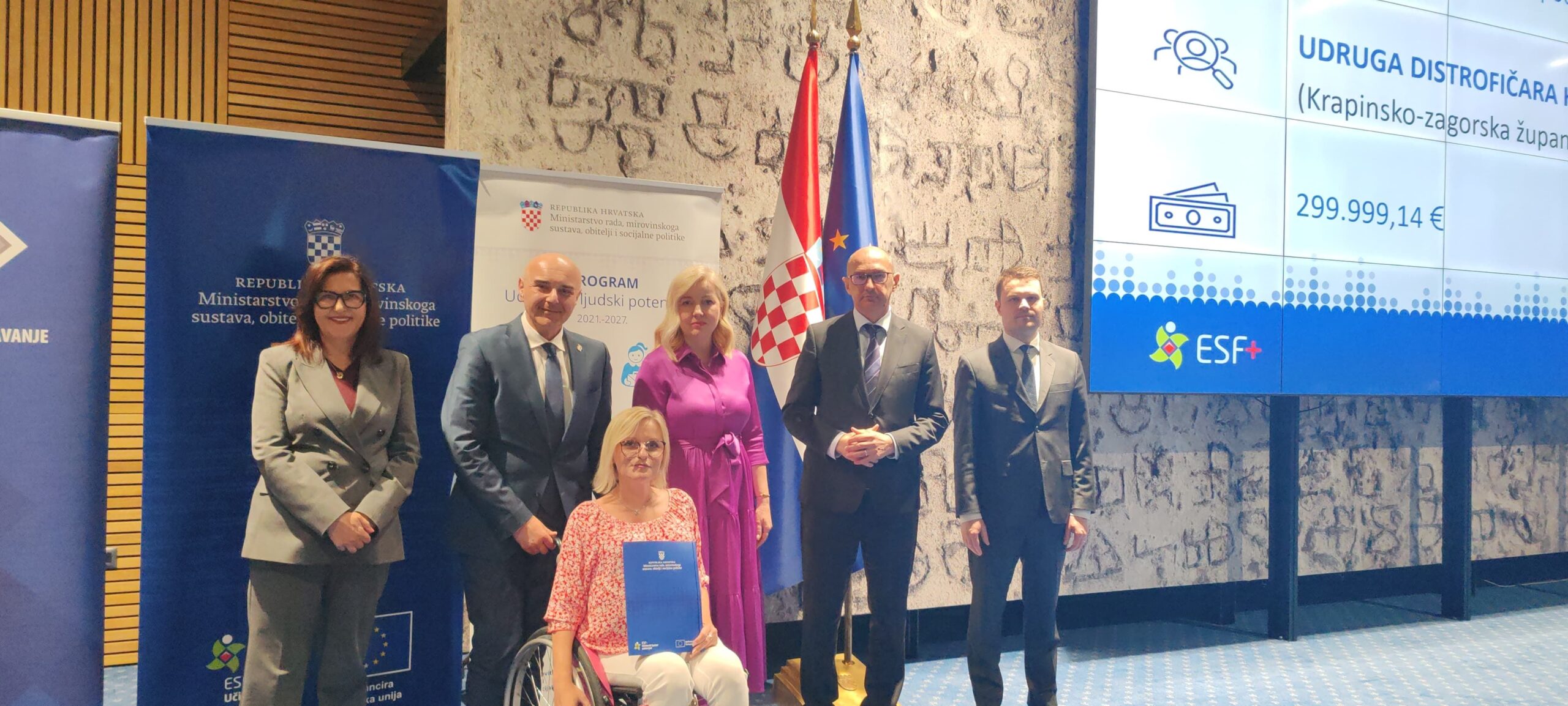 Na fotografiji se nalazi skupina od šest osoba u svečanom prostoru, vjerojatno tijekom službenog događanja ili dodjele ugovora. U prvom planu, u sredini, sjedi žena u invalidskim kolicima koja u rukama drži plavu mapu – najvjerojatnije potpisani ugovor ili projektni dokument. Oko nje stoje četiri muškarca i jedna žena, svi odjeveni u poslovnu, formalnu odjeću, što dodatno naglašava službeni karakter događaja. U pozadini se vide zastave Republike Hrvatske i Europske unije, kao i promotivni paneli s logotipima institucija i fondova. Na velikom ekranu s desne strane prikazan je naziv udruge i iznos projekta (299.999,14 eura), uz oznaku ESF+ fonda. Cjelokupna scena odaje dojam važnog i svečanog trenutka – potpisivanja i dodjele sredstava za projekt koji ima značajnu društvenu vrijednost, posebno u području podrške osobama s invaliditetom.