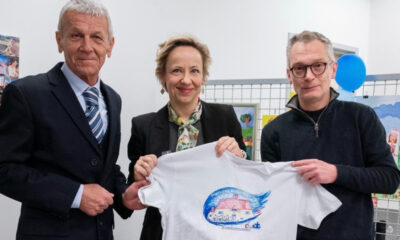 Tri osobe stoje zajedno i drže bijelu majicu s dječjim crtežom, u prostoru s izloženim radovima u pozadini.