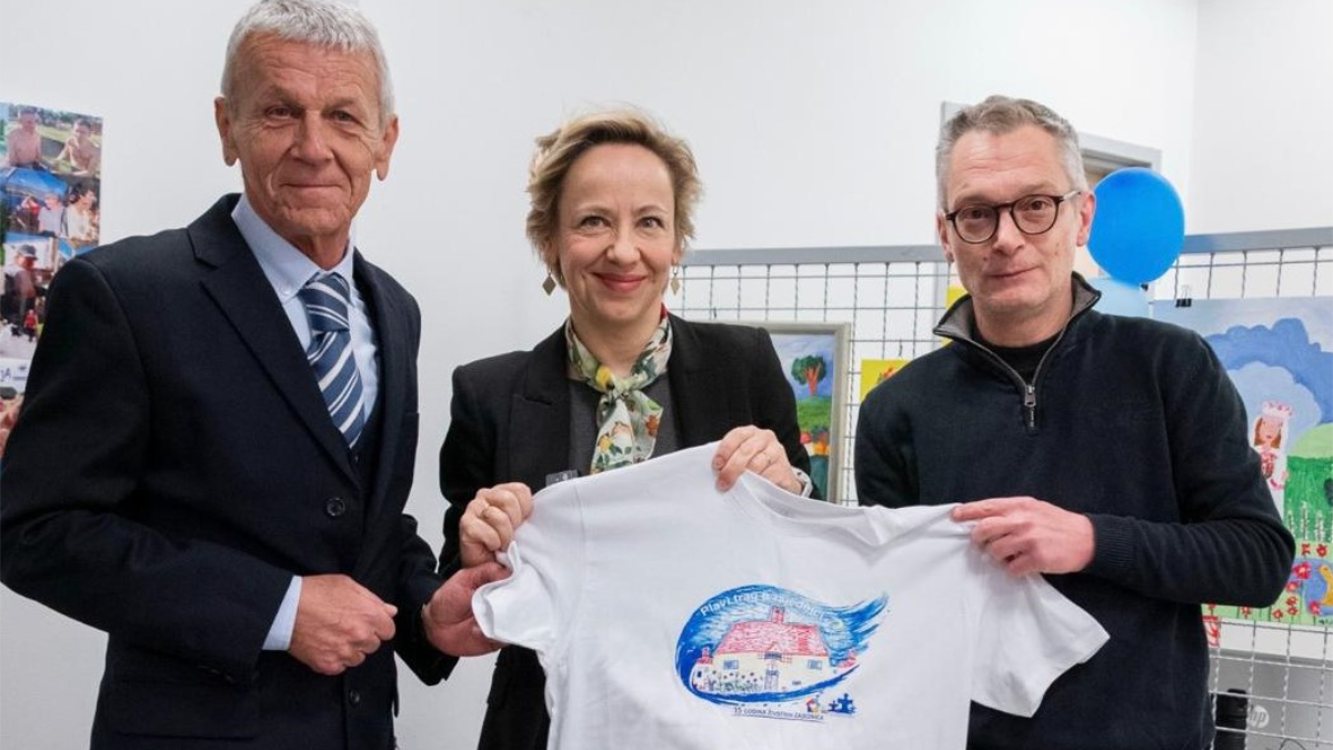 Tri osobe stoje zajedno i drže bijelu majicu s dječjim crtežom, u prostoru s izloženim radovima u pozadini.