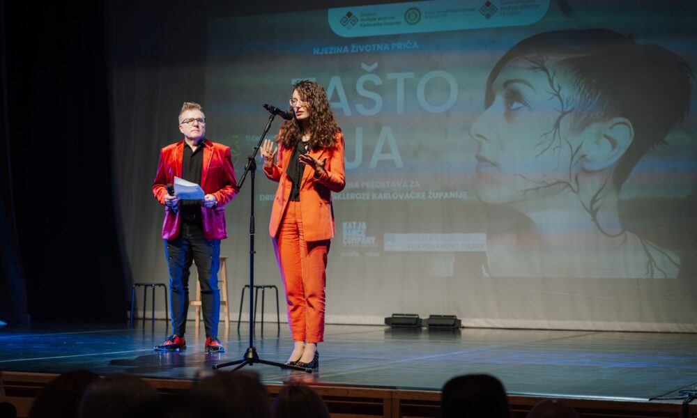 Na fotografiji je prikazan trenutak s pozornice tijekom događaja u kazalištu. U središtu stoji žena odjevena u elegantno narančasto odijelo, koja govori u mikrofon i gestikulira rukama, ostavljajući dojam samouvjerenog i emotivnog obraćanja publici. Pokraj nje, s lijeve strane, stoji muškarac u crvenom sakou koji u rukama drži papire i promatra govor, spreman sudjelovati ili nastaviti program. U pozadini se na velikom projekcijskom platnu vidi vizual predstave s naslovom 'Zašto ja', uz umjetnički prikaz ženskog lica, što dodatno naglašava tematsku ozbiljnost i emocionalnu dubinu događaja. Na pozornici se nalaze i jednostavne stolice, dok se u prvom planu nazire publika koja prati program. Cjelokupna atmosfera fotografije odaje dojam svečanosti, umjetničkog izraza i snažne poruke, s naglaskom na važnost teme i povezanost izvođača s publikom.