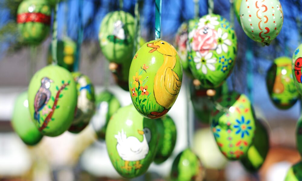Na fotografiji se vide ukrašene uskrsne pisanice obješene na granama stabla. Pisanice su pretežno zelene boje i oslikane raznim motivima poput cvijeća, ptica i životinja. U središtu se ističe jaje s naslikanom žutom ptičicom, dok se u pozadini vide i druga jaja s detaljnim i šarenim uzorcima. Pisanice vise na tankim trakama i lagano se njišu, a pozadina je zamućena, što dodatno naglašava njihovu boju i detalje. Cijela scena djeluje veselo i proljetno, s puno svježih boja i uskrsnog ugođaja.