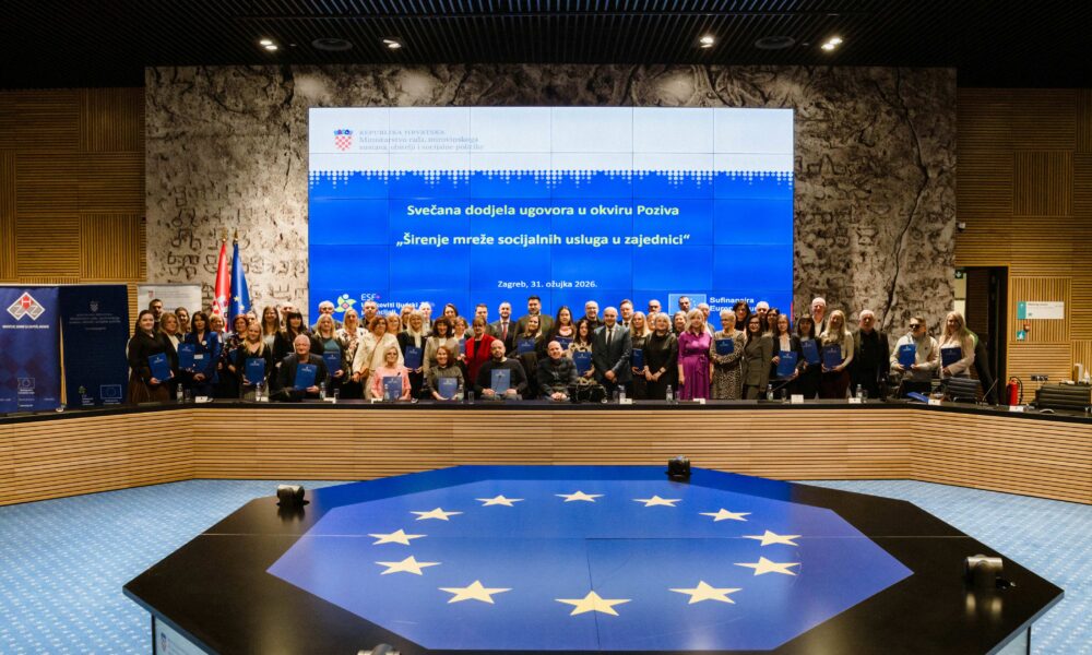 Na slici je prikazana svečana dodjela ugovora održana u velikoj konferencijskoj dvorani. U prvom planu nalazi se veliki stol s motivom zastave Europske unije – plava podloga s krugom žutih zvijezda. U pozadini, na pozornici, stoji velika skupina ljudi – sudionici događaja – koji drže plave mape s ugovorima. Riječ je o predstavnicima različitih organizacija i institucija. Iza njih se nalazi veliki ekran na kojem piše: “Svečana dodjela ugovora u okviru Poziva ‘Širenje mreže socijalnih usluga u zajednici’”, uz datum i mjesto održavanja (Zagreb, 31. ožujka 2026.). S lijeve strane vide se zastave Republike Hrvatske i Europske unije, dok je cijeli prostor moderno uređen, s drvenim i kamenim zidovima te profesionalnom rasvjetom. Atmosfera djeluje formalno i svečano, a fotografija je očito snimljena kao zajednička uspomena nakon potpisivanja ugovora.