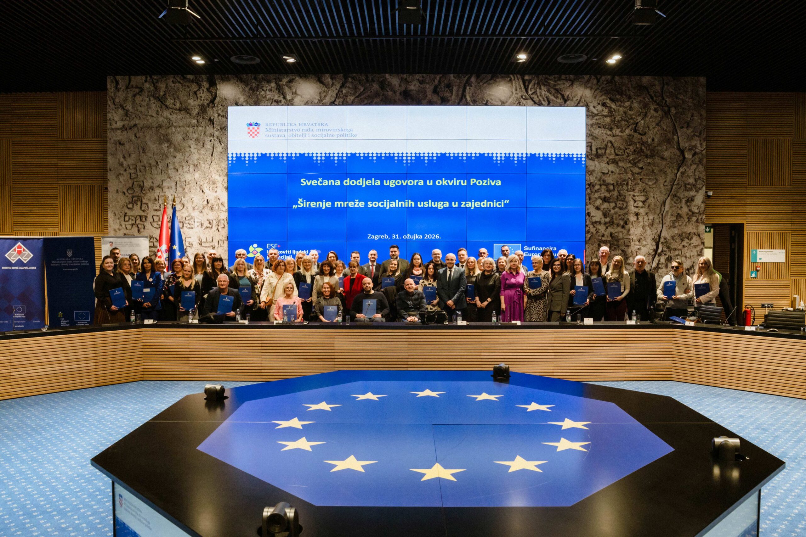 Na slici je prikazana svečana dodjela ugovora održana u velikoj konferencijskoj dvorani. U prvom planu nalazi se veliki stol s motivom zastave Europske unije – plava podloga s krugom žutih zvijezda. U pozadini, na pozornici, stoji velika skupina ljudi – sudionici događaja – koji drže plave mape s ugovorima. Riječ je o predstavnicima različitih organizacija i institucija. Iza njih se nalazi veliki ekran na kojem piše: “Svečana dodjela ugovora u okviru Poziva ‘Širenje mreže socijalnih usluga u zajednici’”, uz datum i mjesto održavanja (Zagreb, 31. ožujka 2026.). S lijeve strane vide se zastave Republike Hrvatske i Europske unije, dok je cijeli prostor moderno uređen, s drvenim i kamenim zidovima te profesionalnom rasvjetom. Atmosfera djeluje formalno i svečano, a fotografija je očito snimljena kao zajednička uspomena nakon potpisivanja ugovora.