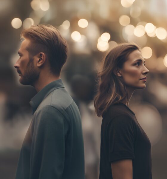 Na fotografiji su prikazani muškarac i žena koji stoje leđima okrenuti jedno drugome, stvarajući snažan dojam emocionalne distance. Oboje gledaju u suprotnim smjerovima, s ozbiljnim i zamišljenim izrazima lica, što sugerira napetost, nesuglasice ili unutarnji konflikt. Pozadina je zamućena i ispunjena toplim, zlatnim svjetlima koja podsjećaju na gradski ambijent ili večernju atmosferu s lampicama, što stvara kontrast između romantičnog okruženja i hladnoće među osobama u prvom planu. Muškarac ima kratku, urednu kosu i bradu te nosi jednostavnu majicu, dok žena ima lagano valovitu kosu i također je odjevena ležerno. Cijela scena odiše tišinom i emocionalnom napetošću, kao da prikazuje trenutak nakon svađe ili razilaženja, gdje su riječi izostale, ali osjećaji ostaju snažno prisutni.