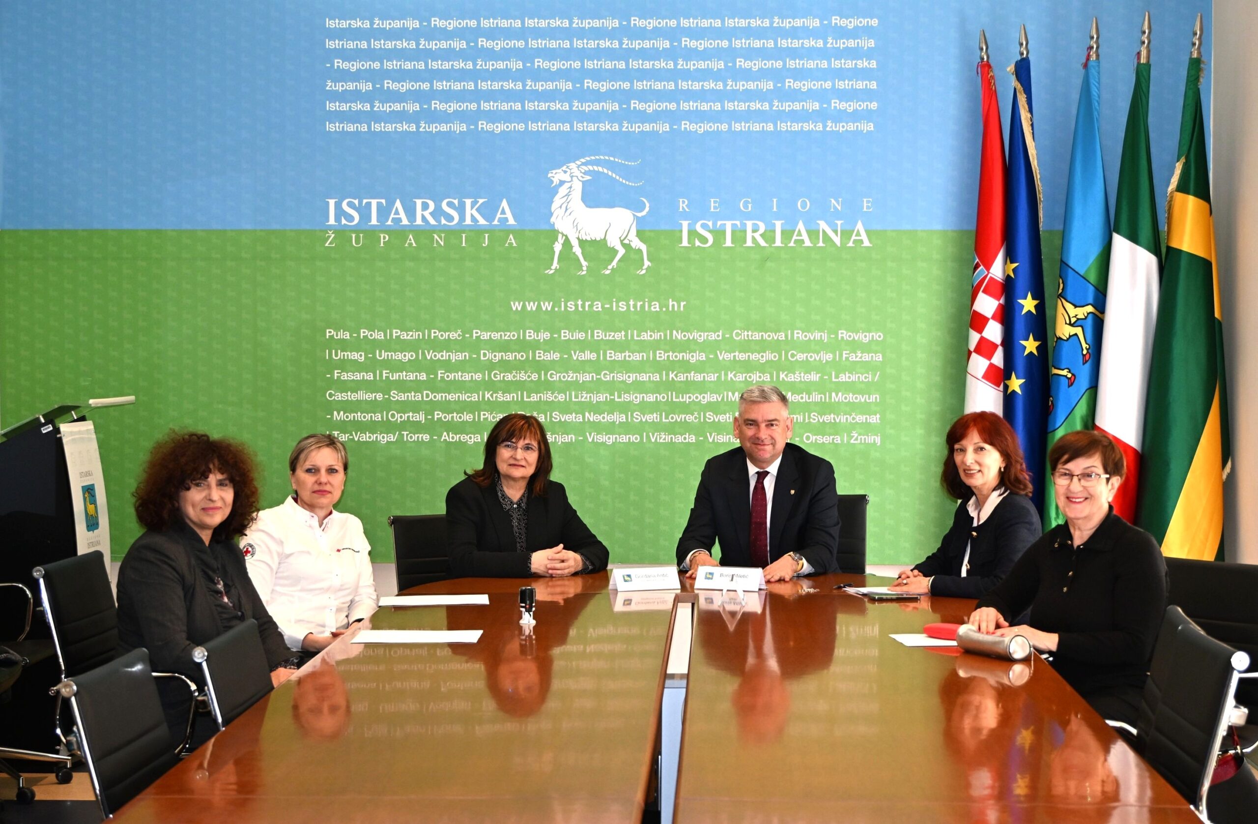 Na fotografiji je prikazan službeni sastanak u prostorijama Istarske županije. Za velikim konferencijskim stolom sjedi šest osoba – jedan muškarac u sredini i pet žena raspoređenih s obje strane stola. Svi su odjeveni poslovno i okrenuti prema kameri, a atmosfera djeluje formalno i profesionalno. U pozadini se nalazi zid s vizualnim identitetom Istarske županije, uključujući naziv na hrvatskom i talijanskom jeziku te prepoznatljiv simbol koze. S desne strane istaknute su zastave – hrvatska zastava, zastava Europske unije te regionalne zastave. Na stolu ispred sudionika nalaze se dokumenti i natpisne pločice s imenima, što upućuje na službeni čin, poput potpisivanja ugovora ili održavanja važnog sastanka. Fotografija odaje dojam institucionalnog događaja vezanog uz javnu upravu ili društvene projekte.