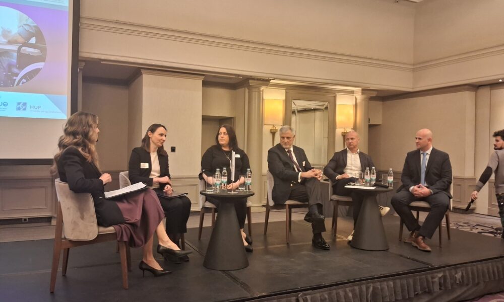 Fotografija prikazuje nekoliko ljudi kako sjedi za stolom i priča, a iza njih se nalazi video zid.