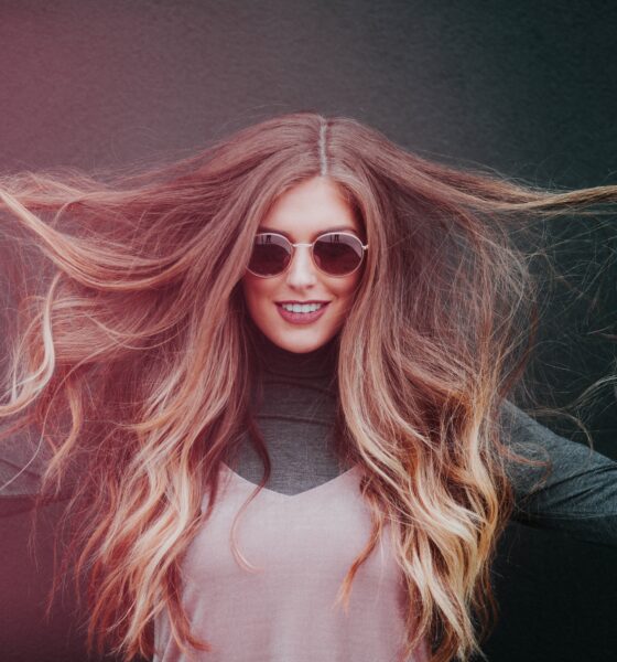 Na slici se nalazi mlada žena s dugom, gustom i valovitom kosom koja se proteže gotovo do struka. Kosa joj je svijetlosmeđa s toplim, zlatnim pramenovima, a razdjeljak je na sredini. Drži pramenove kose u obje ruke i lagano ih podiže u stranu, stvarajući dojam volumena i pokreta. Nosi tamne sunčane naočale i ima blag, samouvjeren osmijeh. Odjevena je u ležernu kombinaciju – dolčevitu tamne boje ispod svjetlijeg topa bez rukava. Pozadina je tamna i neutralna, što dodatno naglašava njezinu kosu i lice. Ukupni dojam fotografije je moderan i stylish, s naglaskom na zdravu i njegovanu kosu.