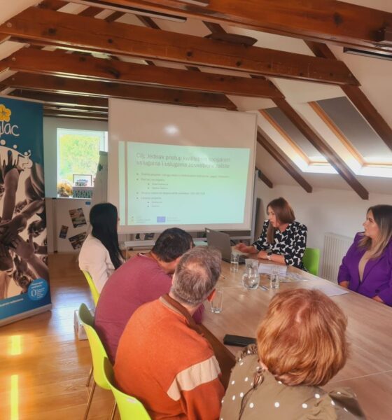 Na slici je prikazan sastanak odnosno konferencija održana u potkrovlju uređenog prostora s drvenim gredama. Za velikim stolom sjedi nekoliko sudionika koji prate prezentaciju prikazanu na platnu putem projektora. U prvom planu vide se sudionici okrenuti prema predavačici koja govori i pokazuje sadržaj prezentacije. S lijeve strane nalazi se promotivni banner Udruge „Jaglac“, dok je na projekcijskom platnu prikazan sadržaj vezan uz socijalne usluge i projektne aktivnosti. Atmosfera djeluje službeno i radno, a konferencija okuplja predstavnike udruge i lokalne zajednice.