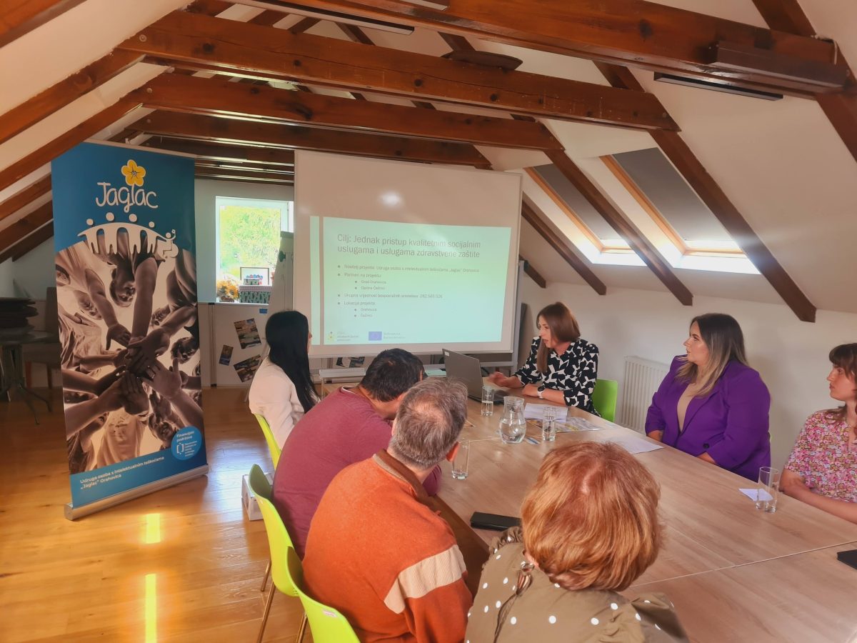 Na slici je prikazan sastanak odnosno konferencija održana u potkrovlju uređenog prostora s drvenim gredama. Za velikim stolom sjedi nekoliko sudionika koji prate prezentaciju prikazanu na platnu putem projektora. U prvom planu vide se sudionici okrenuti prema predavačici koja govori i pokazuje sadržaj prezentacije. S lijeve strane nalazi se promotivni banner Udruge „Jaglac“, dok je na projekcijskom platnu prikazan sadržaj vezan uz socijalne usluge i projektne aktivnosti. Atmosfera djeluje službeno i radno, a konferencija okuplja predstavnike udruge i lokalne zajednice.