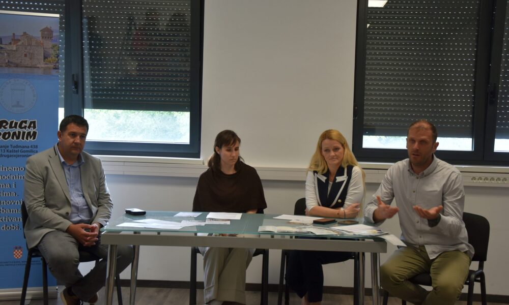 Na fotografiji se nalazi četvero sudionika tijekom predstavljanja projekta ili konferencije za medije u zatvorenom prostoru. Sjede za stolom na kojem su raspoređeni dokumenti i promotivni materijali, dok muškarac s desne strane gestikulacijom objašnjava ili govori okupljenima. U pozadini se vide veliki prozori sa spuštenim roletama te promotivni pano Udruge sv. Jeronim. Atmosfera djeluje formalno i radno, a fotografija prikazuje trenutak predstavljanja projekta vezanog uz socijalne usluge i podršku zajednici.