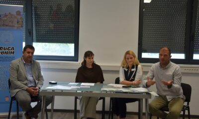 Na fotografiji se nalazi četvero sudionika tijekom predstavljanja projekta ili konferencije za medije u zatvorenom prostoru. Sjede za stolom na kojem su raspoređeni dokumenti i promotivni materijali, dok muškarac s desne strane gestikulacijom objašnjava ili govori okupljenima. U pozadini se vide veliki prozori sa spuštenim roletama te promotivni pano Udruge sv. Jeronim. Atmosfera djeluje formalno i radno, a fotografija prikazuje trenutak predstavljanja projekta vezanog uz socijalne usluge i podršku zajednici.