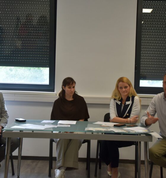 Na fotografiji se nalazi četvero sudionika tijekom predstavljanja projekta ili konferencije za medije u zatvorenom prostoru. Sjede za stolom na kojem su raspoređeni dokumenti i promotivni materijali, dok muškarac s desne strane gestikulacijom objašnjava ili govori okupljenima. U pozadini se vide veliki prozori sa spuštenim roletama te promotivni pano Udruge sv. Jeronim. Atmosfera djeluje formalno i radno, a fotografija prikazuje trenutak predstavljanja projekta vezanog uz socijalne usluge i podršku zajednici.