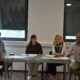 Na fotografiji se nalazi četvero sudionika tijekom predstavljanja projekta ili konferencije za medije u zatvorenom prostoru. Sjede za stolom na kojem su raspoređeni dokumenti i promotivni materijali, dok muškarac s desne strane gestikulacijom objašnjava ili govori okupljenima. U pozadini se vide veliki prozori sa spuštenim roletama te promotivni pano Udruge sv. Jeronim. Atmosfera djeluje formalno i radno, a fotografija prikazuje trenutak predstavljanja projekta vezanog uz socijalne usluge i podršku zajednici.