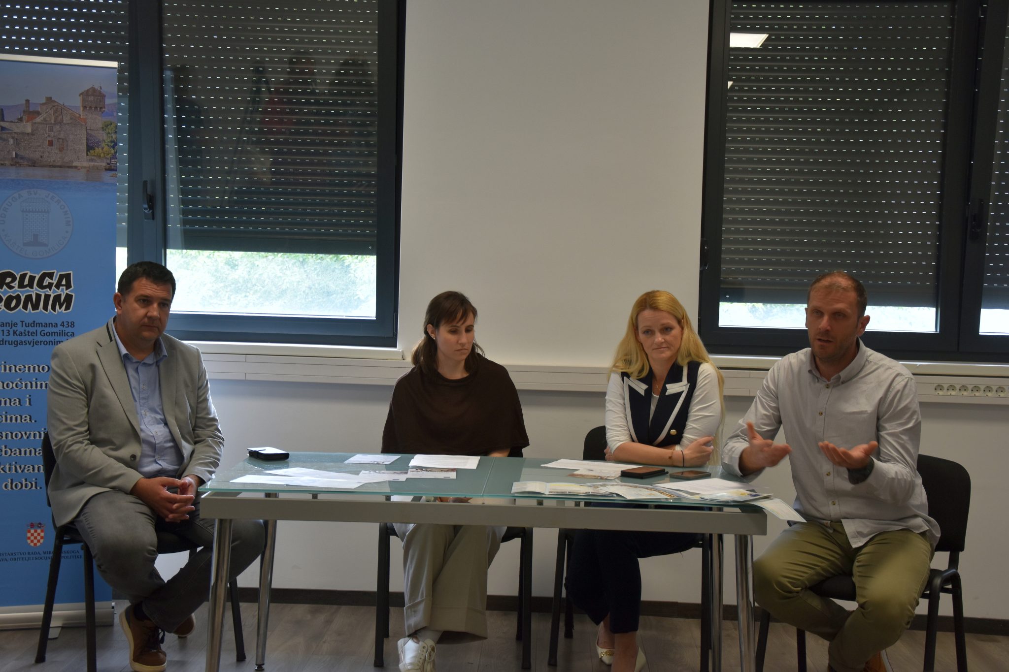 Na fotografiji se nalazi četvero sudionika tijekom predstavljanja projekta ili konferencije za medije u zatvorenom prostoru. Sjede za stolom na kojem su raspoređeni dokumenti i promotivni materijali, dok muškarac s desne strane gestikulacijom objašnjava ili govori okupljenima. U pozadini se vide veliki prozori sa spuštenim roletama te promotivni pano Udruge sv. Jeronim. Atmosfera djeluje formalno i radno, a fotografija prikazuje trenutak predstavljanja projekta vezanog uz socijalne usluge i podršku zajednici.