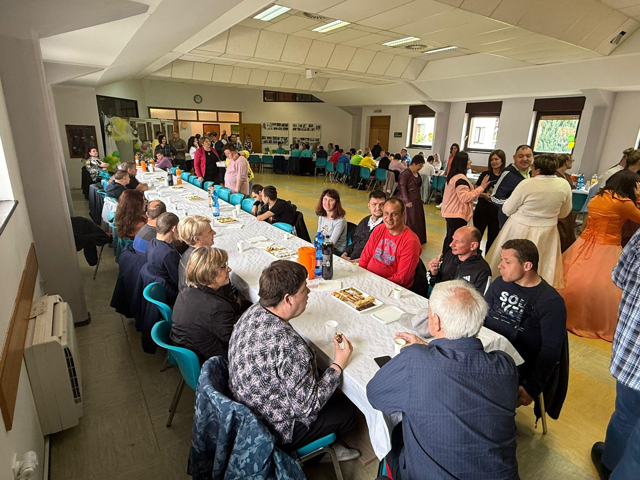 Na fotografiji je prikazan susret udruga osoba s invaliditetom i njihovih korisnika održan u velikoj dvorani Centra Mariapoli u Križevcima. Sudionici sjede za dugim stolovima prekrivenima bijelim stolnjacima, uz druženje, razgovor i osvježenje. U prostoru vlada ugodna i vesela atmosfera, a vidljivo je kako se okupio velik broj ljudi različitih generacija. U pozadini se mogu vidjeti sudionici koji razgovaraju i druže se, dok je dio prostora uređen za zajednički program. Na desnoj strani fotografije nekoliko osoba odjeveno je u svečaniju i scensku odjeću, što upućuje na zabavni dio programa i nastupe sudionika. Fotografija prikazuje toplu i prijateljsku atmosferu zajedništva, međusobnog povezivanja i inkluzije.