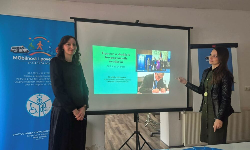 Na fotografiji se nalaze dvije žene u prostoru koji izgleda kao dvorana za prezentacije ili sastanke. Stoje s obje strane projekcijskog platna na kojem je prikazan slajd s naslovom „Ugovor o dodjeli bespovratnih sredstava“ i fotografijama s potpisivanja ugovora. Žena s lijeve strane stoji mirno, odjevena u tamnu odjeću, dok žena s desne strane gestom ruke pokazuje prema projekciji, kao da objašnjava sadržaj prezentacije. Iza njih se nalaze promotivni roll-up plakati s vizualima projekta „Mobilnost i povezanost“, uključujući ilustracije osoba i vozila te logotip udruge. U prvom planu vidi se stol na kojem su uredno posloženi plavi promotivni materijali (vjerojatno brošure ili letci). Prostor djeluje uredno i profesionalno, a cijela scena upućuje na održavanje konferencije ili predstavljanja projekta.