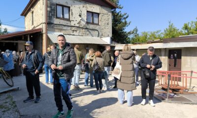 Na fotografiji se nalazi skupina ljudi okupljena ispred stare zgrade koja nosi vidljive tragove ratnih oštećenja. Na pročelju kuće mogu se vidjeti brojna oštećenja i rupe od metaka ili granata, što prostoru daje snažan povijesni i emotivni dojam. Ljudi stoje u manjim grupama, razgovaraju i razgledavaju prostor, dok neki koriste mobitele ili čekaju početak obilaska. U prvom planu nalazi se muškarac u tamnoj jakni i zelenoj trenirci koji stoji okrenut prema fotografu. Oko njega su sudionici izleta odjeveni u ležernu proljetnu odjeću. Vrijeme je sunčano i vedro, a atmosfera djeluje mirno i organizirano, kao dio grupnog posjeta važnoj povijesnoj lokaciji u Sarajevu.