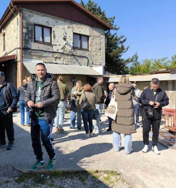 Na fotografiji se nalazi skupina ljudi okupljena ispred stare zgrade koja nosi vidljive tragove ratnih oštećenja. Na pročelju kuće mogu se vidjeti brojna oštećenja i rupe od metaka ili granata, što prostoru daje snažan povijesni i emotivni dojam. Ljudi stoje u manjim grupama, razgovaraju i razgledavaju prostor, dok neki koriste mobitele ili čekaju početak obilaska. U prvom planu nalazi se muškarac u tamnoj jakni i zelenoj trenirci koji stoji okrenut prema fotografu. Oko njega su sudionici izleta odjeveni u ležernu proljetnu odjeću. Vrijeme je sunčano i vedro, a atmosfera djeluje mirno i organizirano, kao dio grupnog posjeta važnoj povijesnoj lokaciji u Sarajevu.
