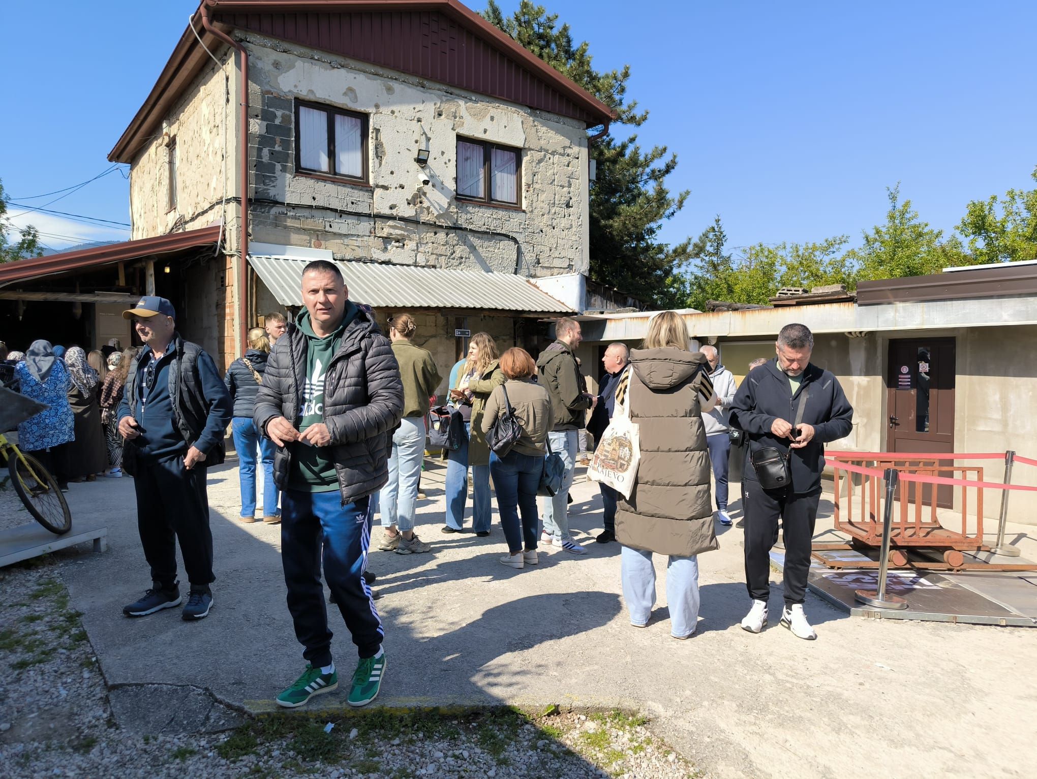 Na fotografiji se nalazi skupina ljudi okupljena ispred stare zgrade koja nosi vidljive tragove ratnih oštećenja. Na pročelju kuće mogu se vidjeti brojna oštećenja i rupe od metaka ili granata, što prostoru daje snažan povijesni i emotivni dojam. Ljudi stoje u manjim grupama, razgovaraju i razgledavaju prostor, dok neki koriste mobitele ili čekaju početak obilaska. U prvom planu nalazi se muškarac u tamnoj jakni i zelenoj trenirci koji stoji okrenut prema fotografu. Oko njega su sudionici izleta odjeveni u ležernu proljetnu odjeću. Vrijeme je sunčano i vedro, a atmosfera djeluje mirno i organizirano, kao dio grupnog posjeta važnoj povijesnoj lokaciji u Sarajevu.