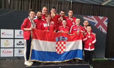 Fotografija prikazuje nekoliko ljudi u crvenim trenirkama s medaljama oko vrata kako drže Hrvatsku zastavu.