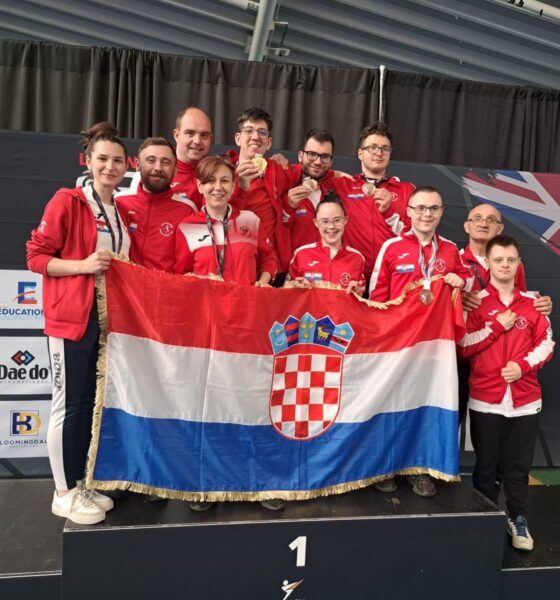 Fotografija prikazuje nekoliko ljudi u crvenim trenirkama s medaljama oko vrata kako drže Hrvatsku zastavu.