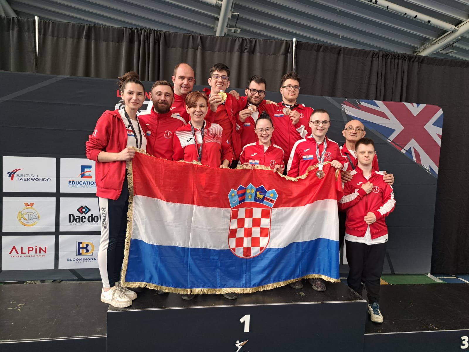 Fotografija prikazuje nekoliko ljudi u crvenim trenirkama s medaljama oko vrata kako drže Hrvatsku zastavu.
