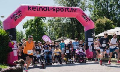 Na fotografiji je prikazan početak humanitarne utrke „Run for MD“ na otvorenom, uz velik broj sudionika različite dobi. Trkači i osobe u invalidskim kolicima zajedno kreću ispod velikog ružičasto-crnog startnog luka s natpisom „keindl-sport.hr“. Događaj se odvija po sunčanom danu u prirodnom okruženju, a uz stazu su okupljeni gledatelji i volonteri. Fotografija prenosi atmosferu zajedništva, inkluzije i podrške osobama s mišićnom distrofijom i drugim neuromuskularnim bolestima.