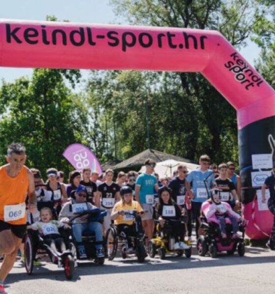 Na fotografiji je prikazan početak humanitarne utrke „Run for MD“ na otvorenom, uz velik broj sudionika različite dobi. Trkači i osobe u invalidskim kolicima zajedno kreću ispod velikog ružičasto-crnog startnog luka s natpisom „keindl-sport.hr“. Događaj se odvija po sunčanom danu u prirodnom okruženju, a uz stazu su okupljeni gledatelji i volonteri. Fotografija prenosi atmosferu zajedništva, inkluzije i podrške osobama s mišićnom distrofijom i drugim neuromuskularnim bolestima.