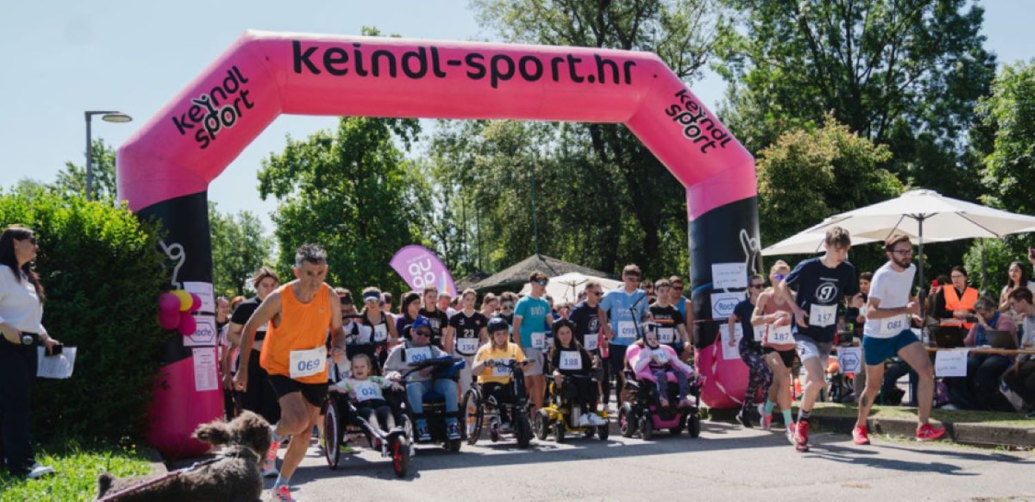 Na fotografiji je prikazan početak humanitarne utrke „Run for MD“ na otvorenom, uz velik broj sudionika različite dobi. Trkači i osobe u invalidskim kolicima zajedno kreću ispod velikog ružičasto-crnog startnog luka s natpisom „keindl-sport.hr“. Događaj se odvija po sunčanom danu u prirodnom okruženju, a uz stazu su okupljeni gledatelji i volonteri. Fotografija prenosi atmosferu zajedništva, inkluzije i podrške osobama s mišićnom distrofijom i drugim neuromuskularnim bolestima.
