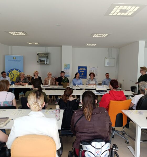Na fotografiji je prikazan skup i radionica održana u svijetloj konferencijskoj prostoriji. Sudionici sjede za stolovima raspoređenima u obliku slova U te pažljivo slušaju govornicu koja stoji s desne strane prostorije i obraća se okupljenima. U prvom planu vide se sudionici okrenuti prema panelu predavača, među kojima su i osobe koje koriste invalidska kolica. Za glavnim stolom sjedi nekoliko sudionika i predstavnika organizacija, a iza njih se nalazi roll-up plakat SOIH-a i promotivni materijali povezani s pravima osoba s invaliditetom i neovisnim življenjem. Atmosfera djeluje radno, ali ugodno i uključivo, s naglaskom na razgovor, suradnju i razmjenu iskustava među sudionicima.