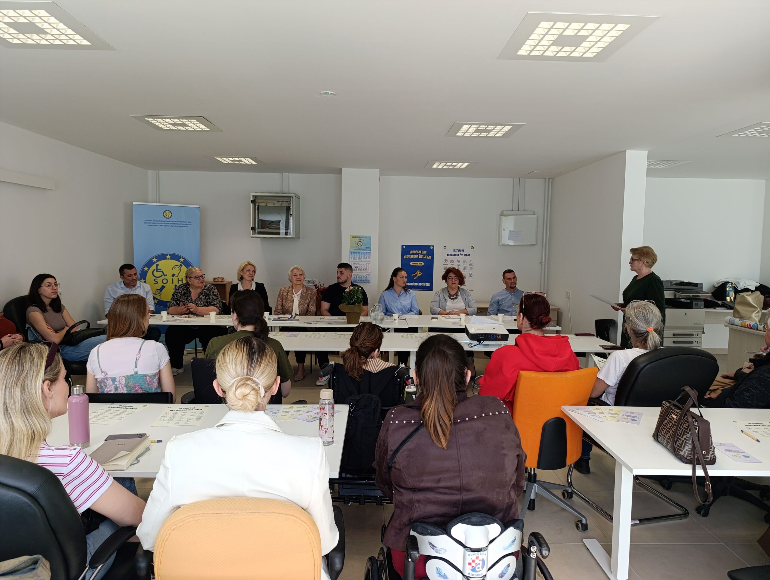 Na fotografiji je prikazan skup i radionica održana u svijetloj konferencijskoj prostoriji. Sudionici sjede za stolovima raspoređenima u obliku slova U te pažljivo slušaju govornicu koja stoji s desne strane prostorije i obraća se okupljenima. U prvom planu vide se sudionici okrenuti prema panelu predavača, među kojima su i osobe koje koriste invalidska kolica. Za glavnim stolom sjedi nekoliko sudionika i predstavnika organizacija, a iza njih se nalazi roll-up plakat SOIH-a i promotivni materijali povezani s pravima osoba s invaliditetom i neovisnim življenjem. Atmosfera djeluje radno, ali ugodno i uključivo, s naglaskom na razgovor, suradnju i razmjenu iskustava među sudionicima.