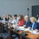 Na fotografiji je prikazan sastanak za dugim konferencijskim stolom, vjerojatno u službenom ili institucionalnom prostoru poput saborske dvorane ili prostorije za sjednice. Za stolom sjedi veći broj sudionika, među kojima su žene i muškarci, odjeveni uglavnom poslovno, što upućuje na formalni karakter događaja. Sudionici imaju ispred sebe mikrofone, dokumente, bilježnice i boce vode, a neki zapisuju bilješke ili pažljivo slušaju govornike. Atmosfera djeluje ozbiljno i koncentrirano, kao tijekom rasprave ili tematske sjednice. U pozadini se vidi kamera na stativu, što sugerira da se događaj snima ili dokumentira. Cjelokupni dojam fotografije upućuje na službenu raspravu ili sastanak stručnjaka i predstavnika institucija.