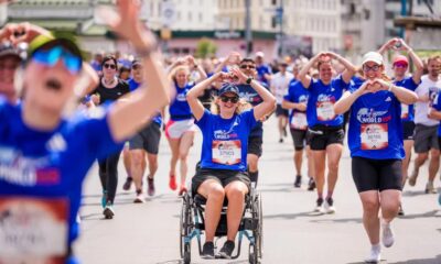 Na fotografiji se vidi velika skupina sudionika tijekom humanitarne utrke Wings for Life World Run. U prvom planu nalazi se djevojka u invalidskim kolicima odjevena u plavu službenu majicu utrke, dok oko nje trče brojni sudionici. Mnogi od njih rukama oblikuju srce, stvarajući toplu i emotivnu atmosferu zajedništva i podrške. Utrka se odvija gradskim ulicama po sunčanom danu, a sudionici djeluju nasmijano i motivirano. Fotografija snažno prenosi poruku inkluzije, humanosti i zajedničkog cilja – pomoći istraživanju ozljeda leđne moždine.