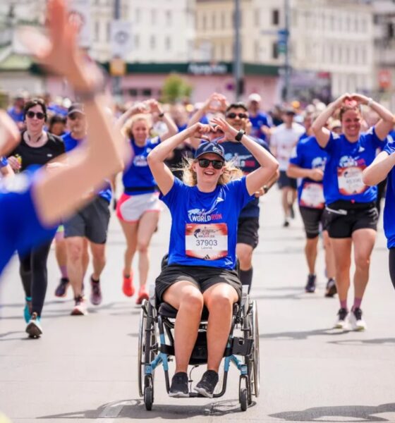 Na fotografiji se vidi velika skupina sudionika tijekom humanitarne utrke Wings for Life World Run. U prvom planu nalazi se djevojka u invalidskim kolicima odjevena u plavu službenu majicu utrke, dok oko nje trče brojni sudionici. Mnogi od njih rukama oblikuju srce, stvarajući toplu i emotivnu atmosferu zajedništva i podrške. Utrka se odvija gradskim ulicama po sunčanom danu, a sudionici djeluju nasmijano i motivirano. Fotografija snažno prenosi poruku inkluzije, humanosti i zajedničkog cilja – pomoći istraživanju ozljeda leđne moždine.