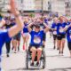 Na fotografiji se vidi velika skupina sudionika tijekom humanitarne utrke Wings for Life World Run. U prvom planu nalazi se djevojka u invalidskim kolicima odjevena u plavu službenu majicu utrke, dok oko nje trče brojni sudionici. Mnogi od njih rukama oblikuju srce, stvarajući toplu i emotivnu atmosferu zajedništva i podrške. Utrka se odvija gradskim ulicama po sunčanom danu, a sudionici djeluju nasmijano i motivirano. Fotografija snažno prenosi poruku inkluzije, humanosti i zajedničkog cilja – pomoći istraživanju ozljeda leđne moždine.