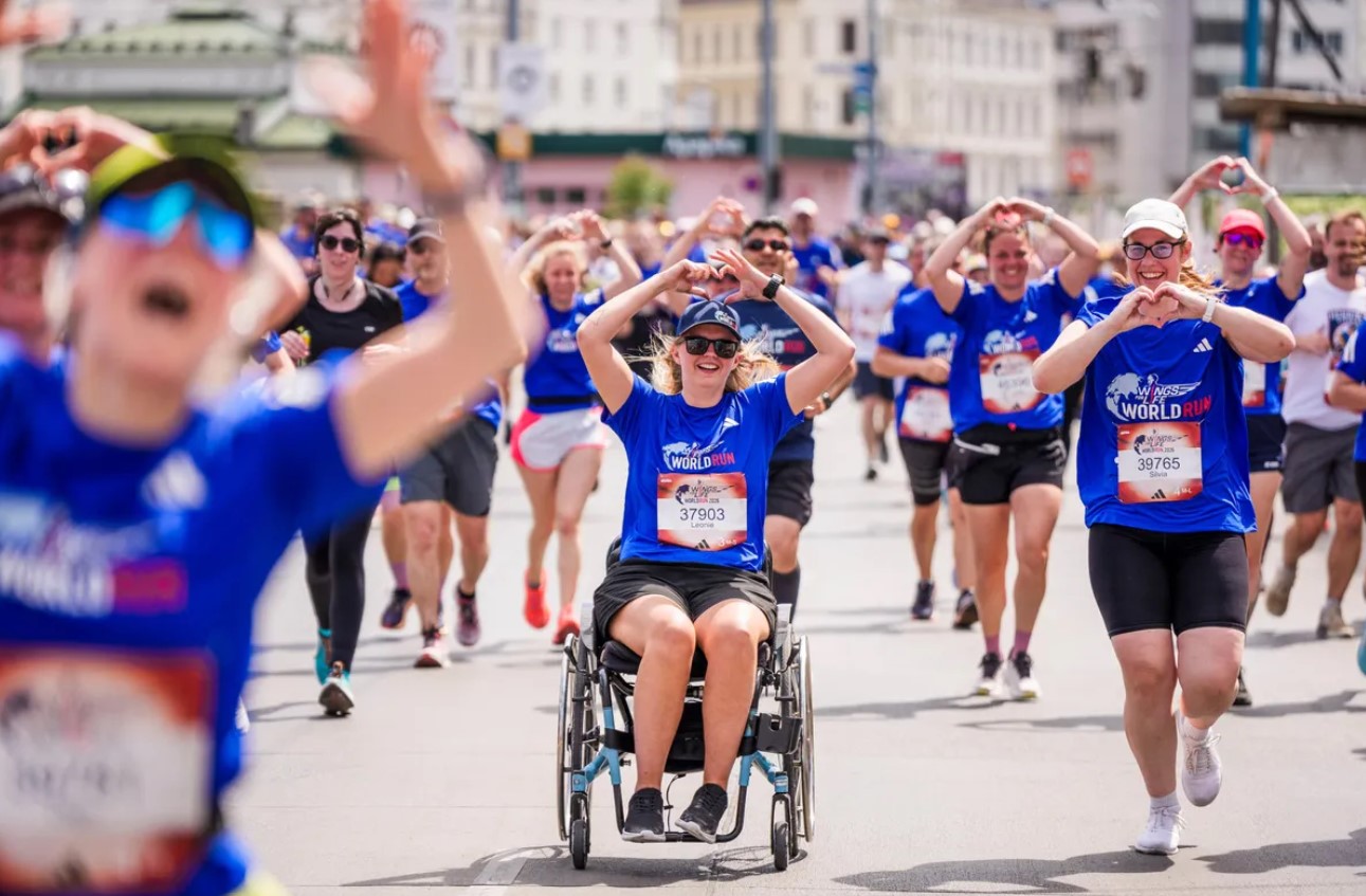 Na fotografiji se vidi velika skupina sudionika tijekom humanitarne utrke Wings for Life World Run. U prvom planu nalazi se djevojka u invalidskim kolicima odjevena u plavu službenu majicu utrke, dok oko nje trče brojni sudionici. Mnogi od njih rukama oblikuju srce, stvarajući toplu i emotivnu atmosferu zajedništva i podrške. Utrka se odvija gradskim ulicama po sunčanom danu, a sudionici djeluju nasmijano i motivirano. Fotografija snažno prenosi poruku inkluzije, humanosti i zajedničkog cilja – pomoći istraživanju ozljeda leđne moždine.