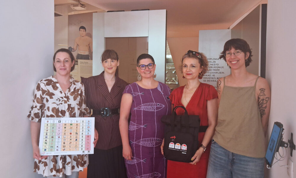 Pet žena pozira u izložbenom prostoru držeći komunikacijsko pomagalo i promotivne materijale.