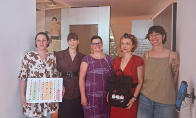 Pet žena pozira u izložbenom prostoru držeći komunikacijsko pomagalo i promotivne materijale.