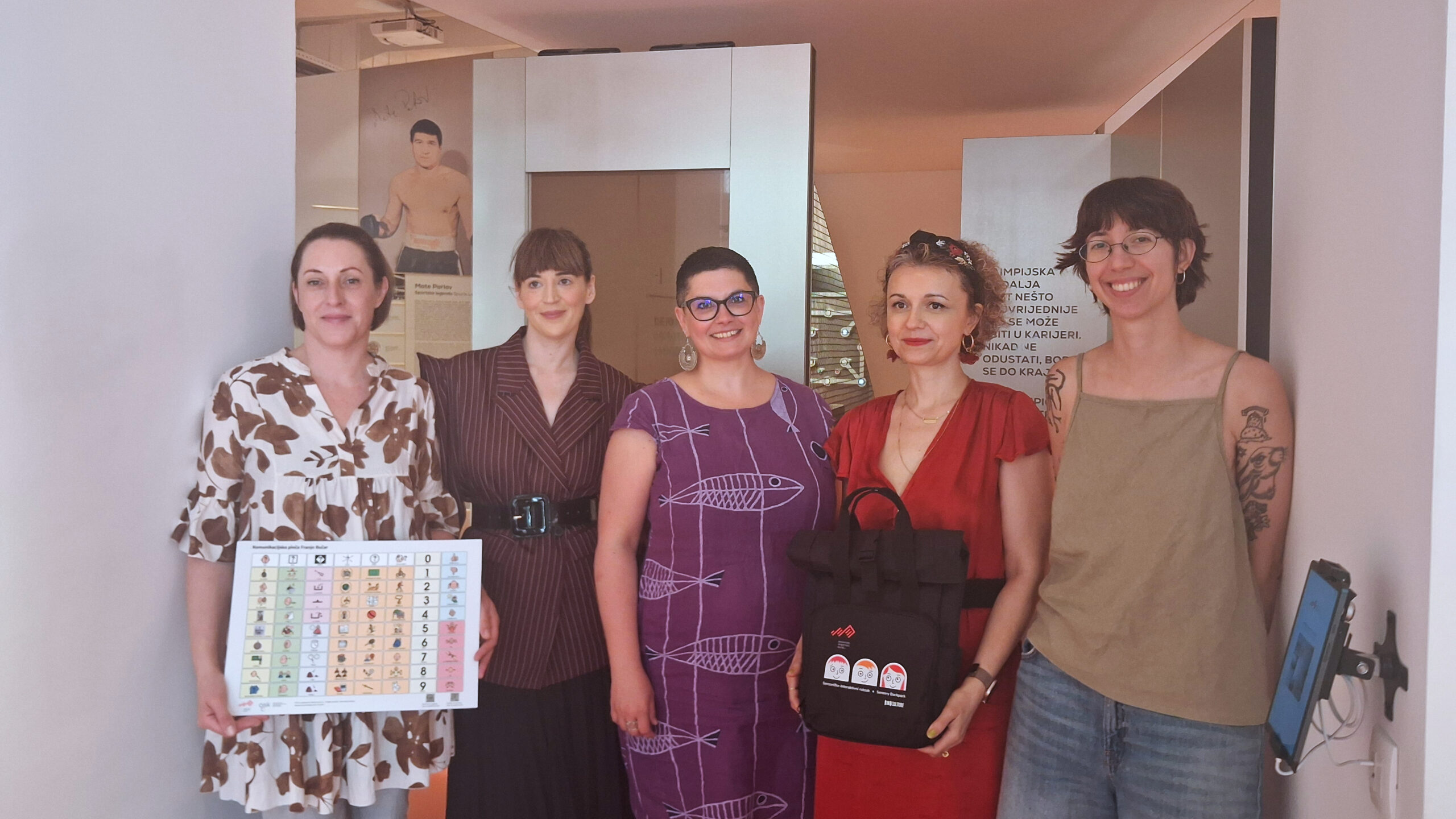 Pet žena pozira u izložbenom prostoru držeći komunikacijsko pomagalo i promotivne materijale.