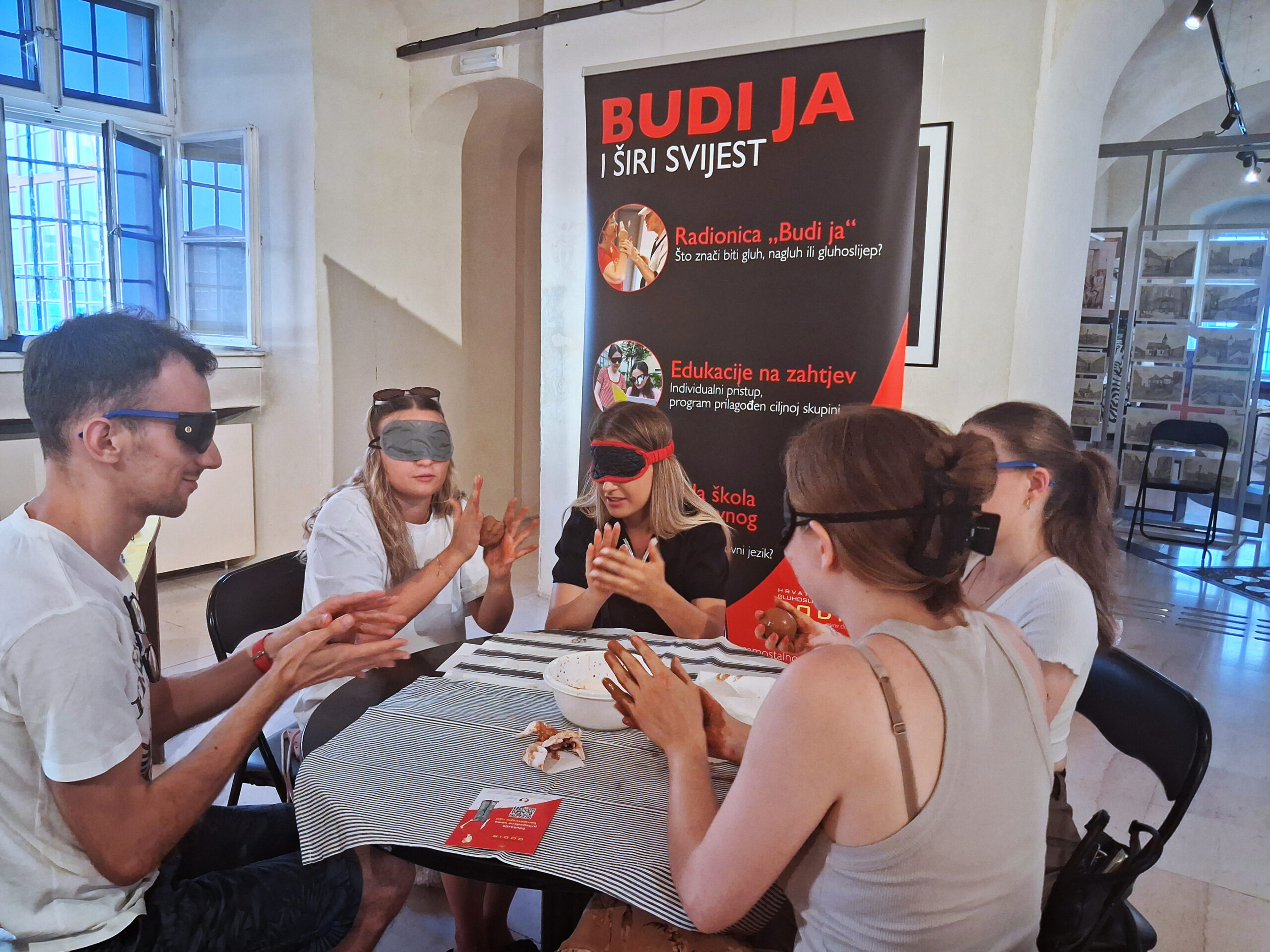 Šest sudionika sjedi za stolom i sudjeluje u interaktivnoj radionici s povezima preko očiju. Ispred njih su zdjele i predmeti koje istražuju dodirom, dok se u pozadini nalazi promotivni pano "Budi ja i širi svijest" s informacijama o edukacijama Hrvatskog saveza gluhoslijepih osoba DODIR.