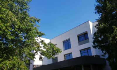 Na fotografiji je prikazana moderna, suvremeno uređena zgrada okružena zelenilom. Objekt ima tri etaže, ravne linije i minimalistički arhitektonski stil, s bijelom fasadom i velikim tamnim staklenim površinama koje prostoru daju moderan izgled. U prizemlju se ističe natkriveni ulaz s tamnom konstrukcijom koja stvara kontrast svijetloj vanjštini zgrade. S obje strane zgrade nalaze se visoka stabla bogatih zelenih krošnji koja djelomično uokviruju kadar i stvaraju ugodan prirodni ambijent. Iznad objekta prostire se vedro, intenzivno plavo nebo bez oblaka, što fotografiji daje svježinu i vedrinu te dodatno naglašava svijetle tonove pročelja. Fotografija ostavlja dojam urednog, funkcionalnog i suvremenog prostora smještenog u mirnom okruženju, u kojem su arhitektura i priroda skladno povezane.