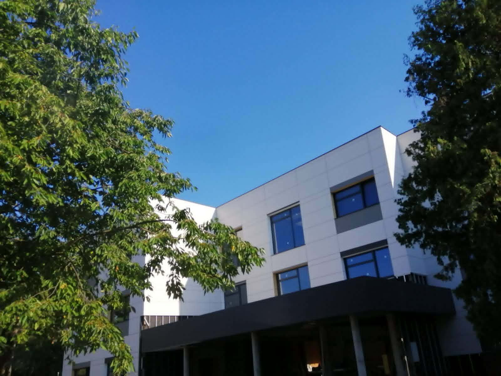 Na fotografiji je prikazana moderna, suvremeno uređena zgrada okružena zelenilom. Objekt ima tri etaže, ravne linije i minimalistički arhitektonski stil, s bijelom fasadom i velikim tamnim staklenim površinama koje prostoru daju moderan izgled. U prizemlju se ističe natkriveni ulaz s tamnom konstrukcijom koja stvara kontrast svijetloj vanjštini zgrade. S obje strane zgrade nalaze se visoka stabla bogatih zelenih krošnji koja djelomično uokviruju kadar i stvaraju ugodan prirodni ambijent. Iznad objekta prostire se vedro, intenzivno plavo nebo bez oblaka, što fotografiji daje svježinu i vedrinu te dodatno naglašava svijetle tonove pročelja. Fotografija ostavlja dojam urednog, funkcionalnog i suvremenog prostora smještenog u mirnom okruženju, u kojem su arhitektura i priroda skladno povezane.