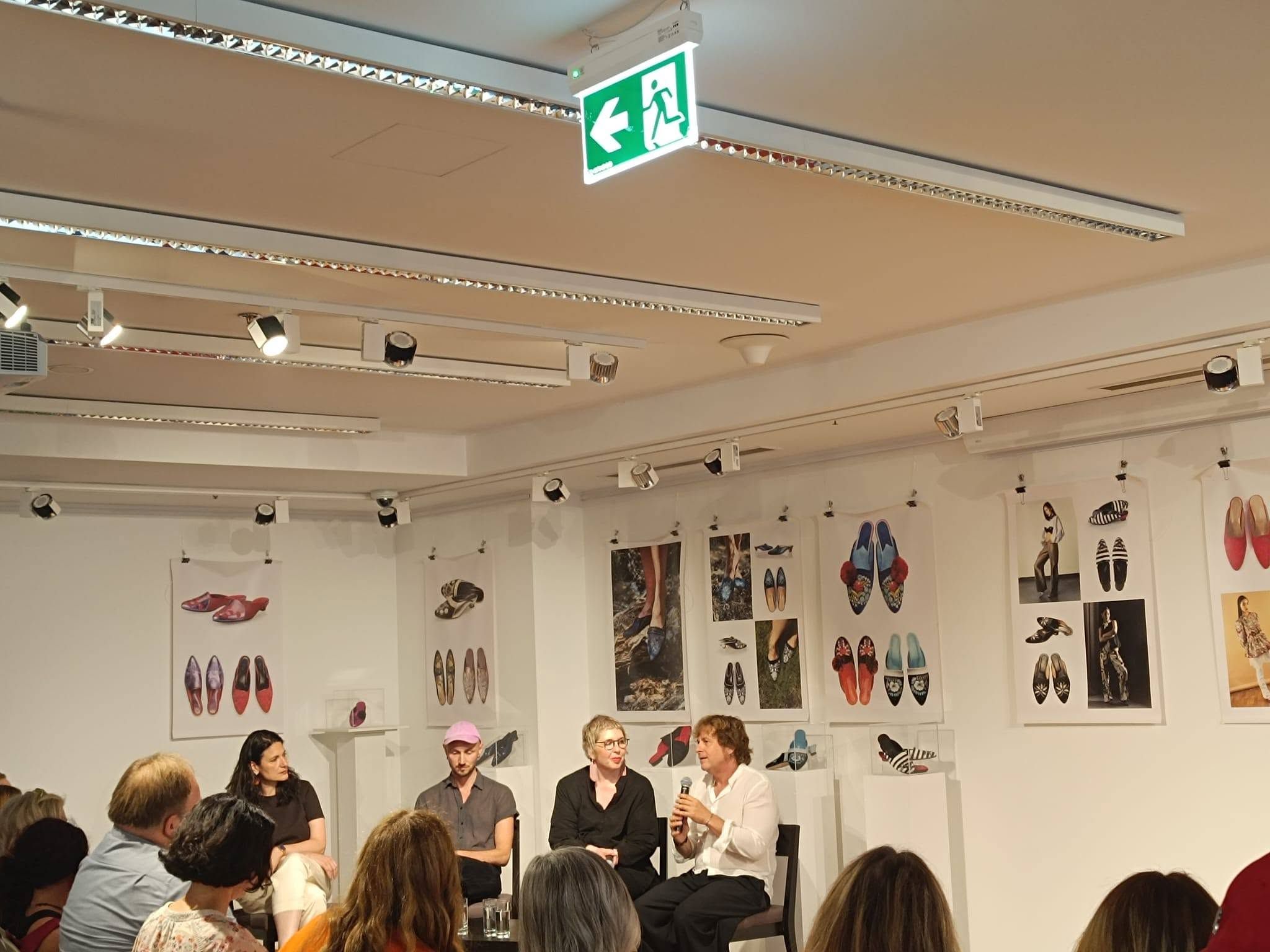 a fotografiji je prikazan razgovor nakon projekcije dokumentarnog filma u izložbenom prostoru kulturnog centra. U prvom planu nalazi se publika koja pažljivo prati panel-raspravu, dok na pozornici sjede četiri sudionika događanja. Jedna od govornica drži mikrofon i obraća se okupljenima, dok ostali sudionici pozorno slušaju. Prostor je uređen kao galerija, a zidove krase izložbeni panoi s fotografijama i umjetničkim prikazima obuće, što dodatno doprinosi umjetničkom ozračju događanja. Atmosfera djeluje ugodno, otvoreno i poticajno za razmjenu ideja, a velik odaziv publike svjedoči o interesu za teme inkluzivne umjetnosti i kulturne suradnje.