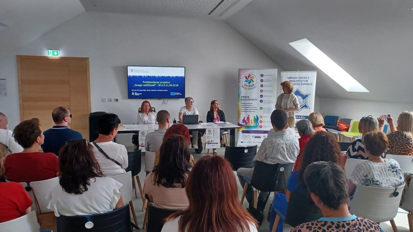 Na fotografiji je prikazan trenutak s početne konferencije projekta „Snaga različitosti“, održane u Multimedijskoj dvorani Kina Edison u Karlovcu. U prvom planu vidi se publika okupljena u dvorani, dok se u pozadini nalaze sudionice panela koje predstavljaju projekt i njegove aktivnosti. Jedna od govornica obraća se prisutnima, a iza nje su postavljeni promotivni roll-up plakati i ekran s nazivom projekta. Atmosfera djeluje radno i svečano, uz velik interes sudionika za teme socijalne uključenosti, podrške osobama s invaliditetom i razvoja socijalnih usluga u zajednici.
