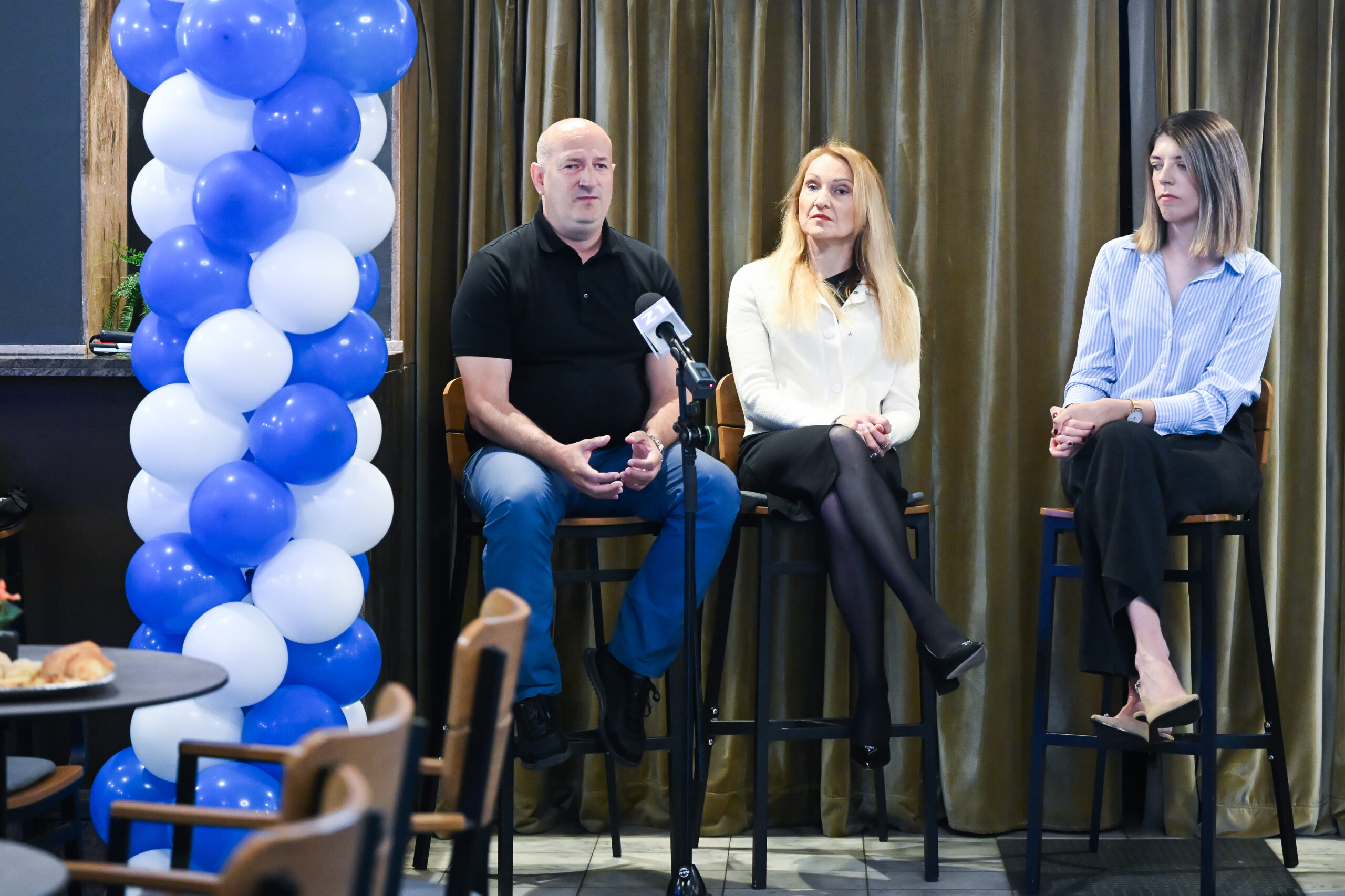 Panel-rasprava s troje sudionika ispred mikrofona i plavo-bijele dekoracije od balona.