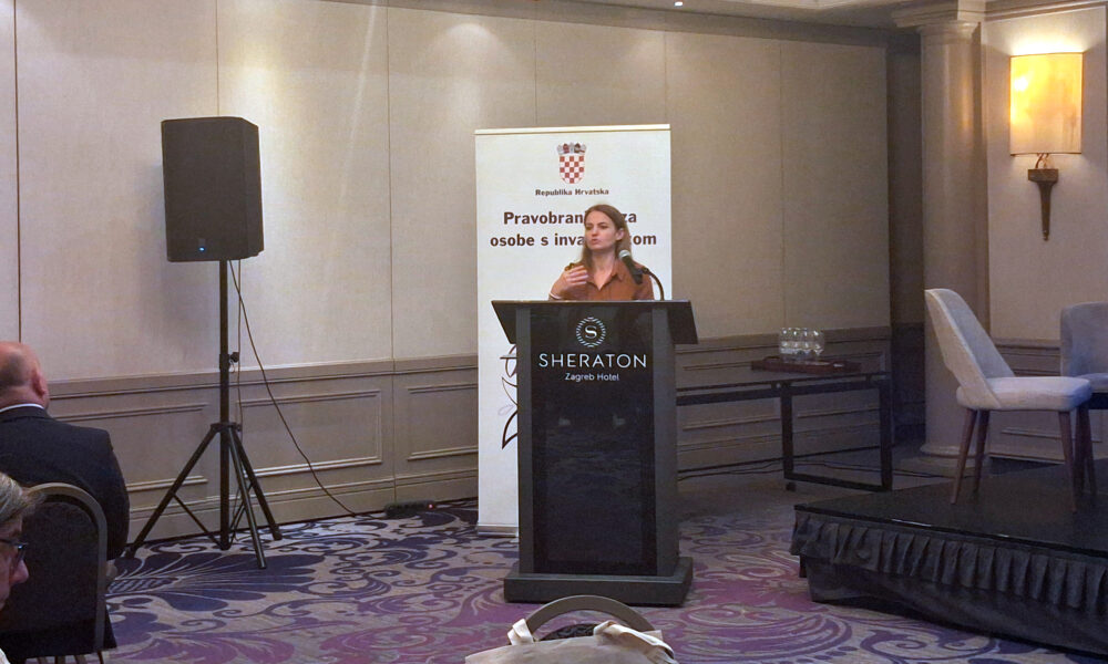 Žena govori za govornicom na konferenciji, ispred bannera Pravobranitelja za osobe s invaliditetom u hotelu Sheraton.