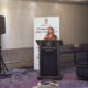 Žena govori za govornicom na konferenciji, ispred bannera Pravobranitelja za osobe s invaliditetom u hotelu Sheraton.